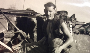 Bill Clevenger, holding his helmet, posed by a jeep in Vietnam.