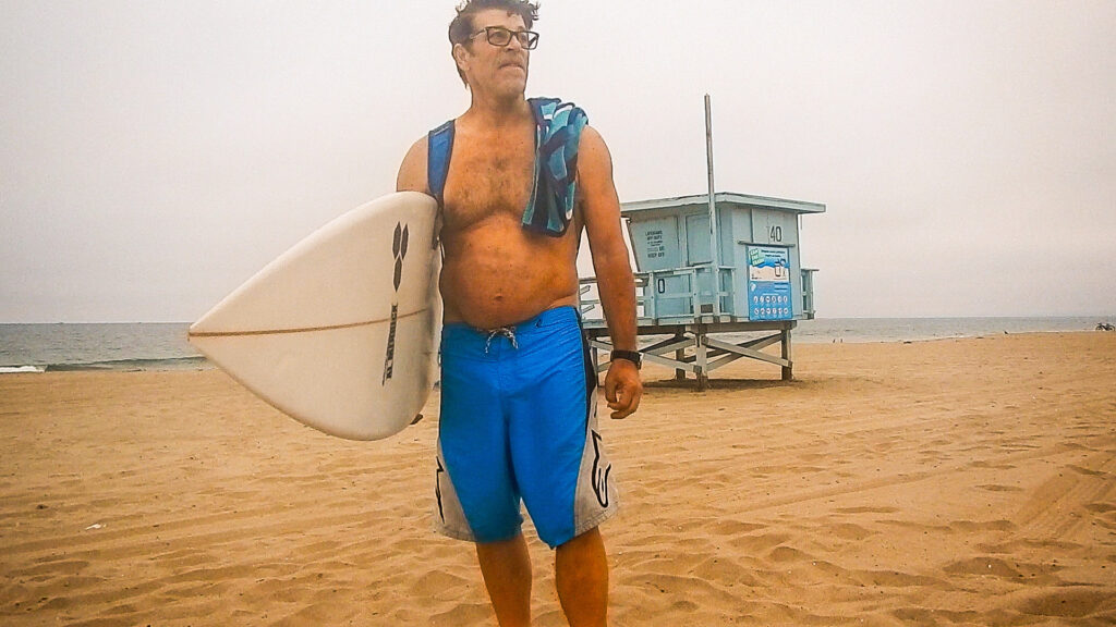 Paul, holding a surfboard, standing on the beach