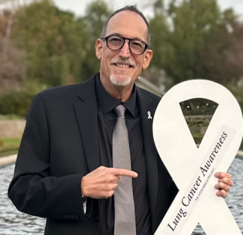 Jeff Stibelman, looking sharp in a black shirt and tie, pointing at the Lung Cancer Awareness Ribbon he's holding