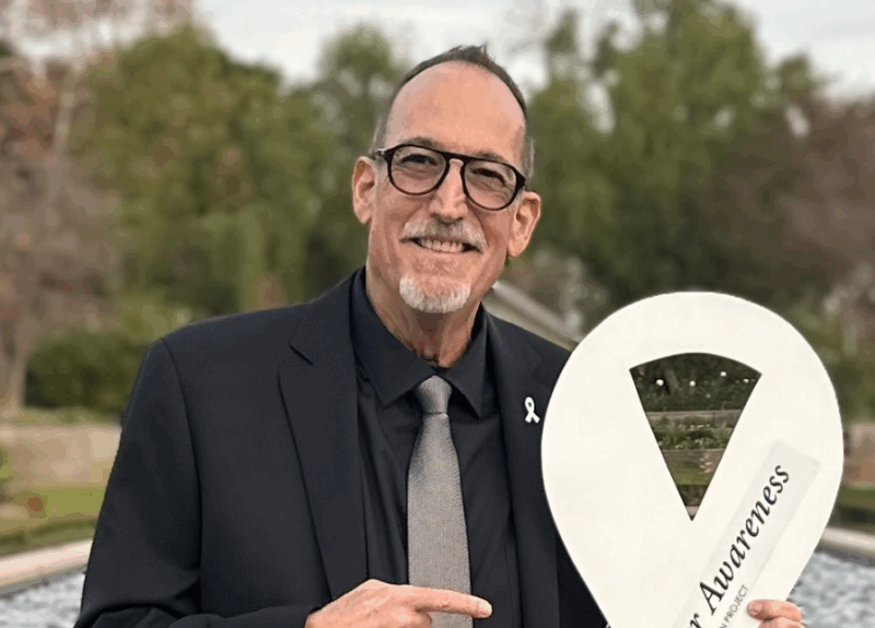 Jeff Stibelman, looking sharp in a black shirt and tie, pointing at the Lung Cancer Awareness Ribbon he's holding